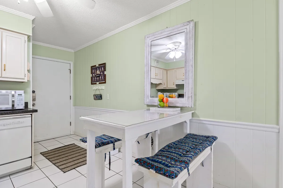 A cheerful dining area with a bright white table, colorful fruit reflected in a mirror, decorative wall art, and blue patterned stools. The space is well-lit and arranged for casual meals or group activities, exuding a relaxed, vacation vibe.
