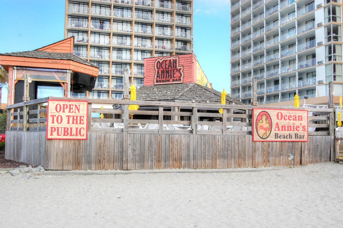 An exterior image of the popular Ocean Annie's bar, featuring a wooden beachside structure with festive yellow umbrellas and signage welcoming guests. The scene suggests an atmosphere of live music, beach parties, and casual drinks, right by the ocean.
