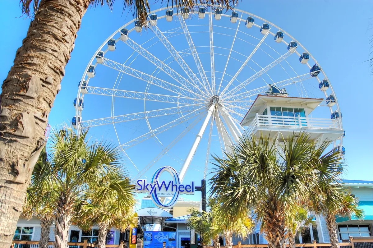 The image spotlights the iconic SkyWheel, surrounded by palm trees and the welcoming entrance building. Bright sunlight sets a joyful mood, and the SkyWheel dominates the skyline, promising fun and breathtaking views for families.