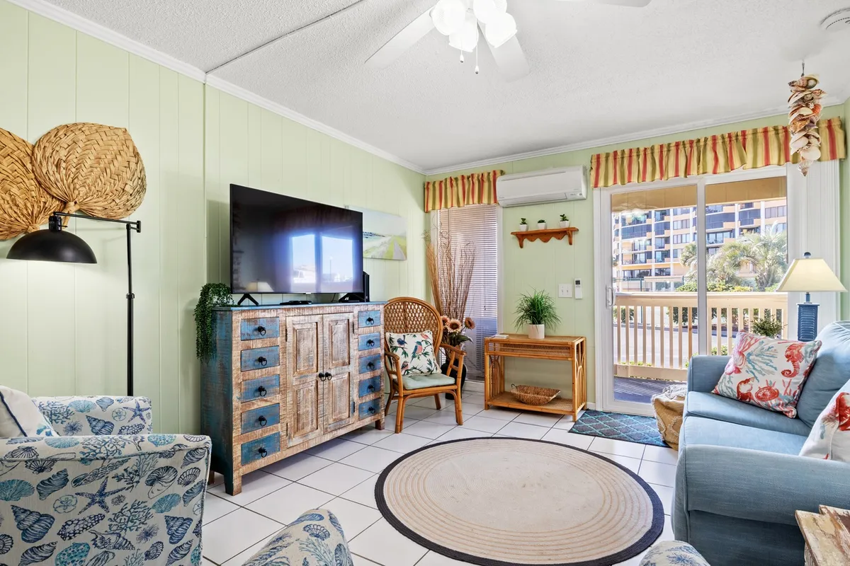 This image features a cozy, open living area with a blue sofa, colorful patterned chairs, a wooden cabinet, a TV, and a large window flooding the space with natural light. The arrangement encourages relaxation and conversation, while the window hints at a connection to the outdoors.