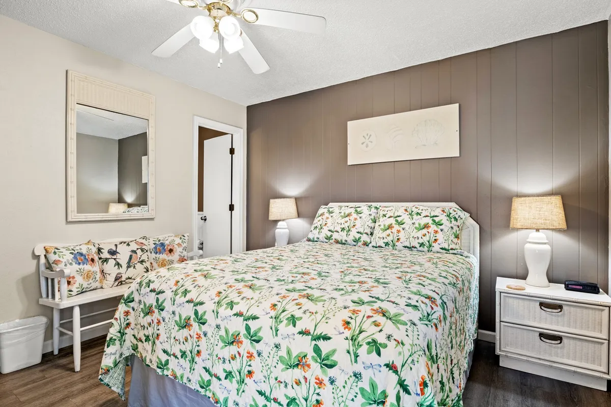 A tranquil bedroom featuring a queen bed adorned with fresh floral bedding, paired with matching nightstands and softly glowing lamps. A decorative mirror and a cushioned bench add both style and comfort, while natural light streams in, highlighting the calm, inviting atmosphere.