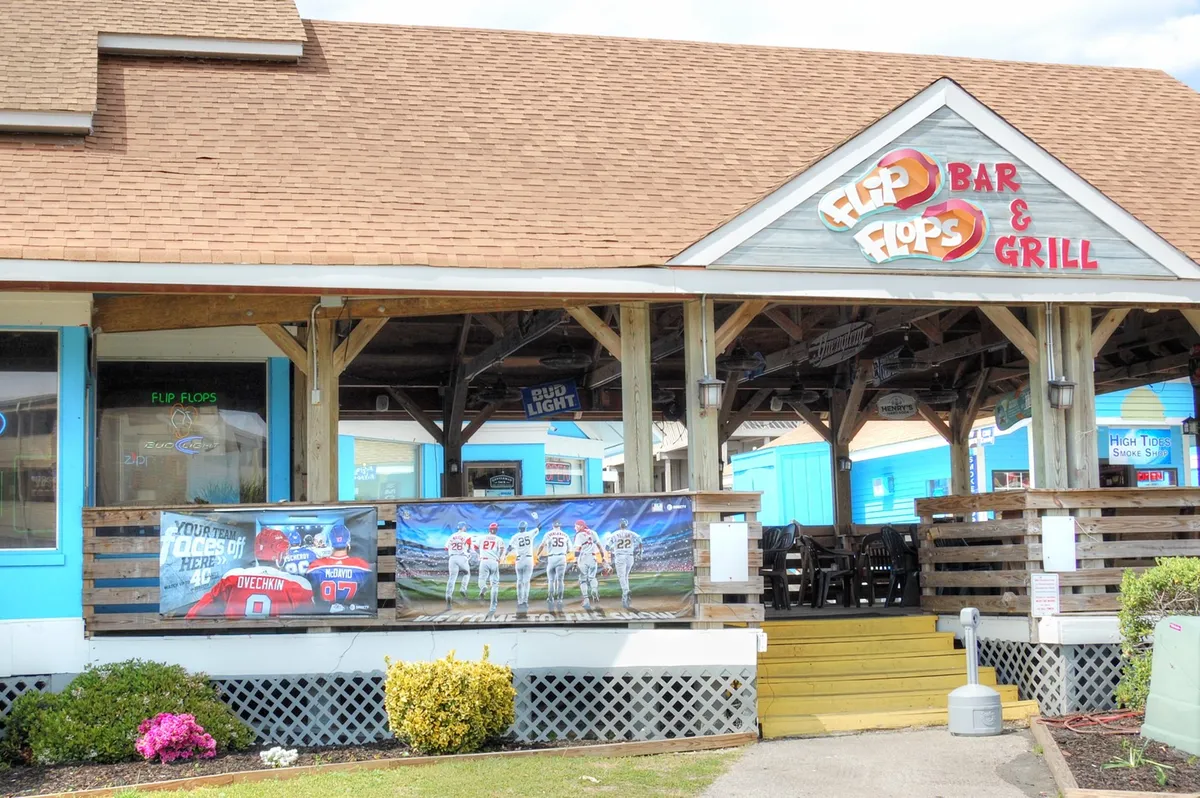 This exterior shot captures the 'Flip Flops Bar & Grill' sign, a vibrant and inviting local venue. The image showcases a lively, colorful building with outdoor seating and banners that hint at sports-themed evenings. The casual, fun atmosphere is apparent, with a street-side view that feels accessible and social.