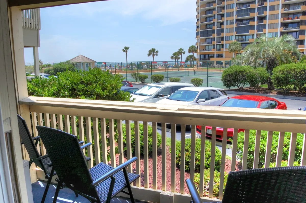 A welcoming balcony with comfortable seating overlooks the tennis courts, swaying palm trees, and the shimmering ocean beyond. The relaxing scene is bathed in warm natural light, inviting leisurely moments spent outdoors. The setting is tranquil and ideal for morning coffee or unwinding at sunset.