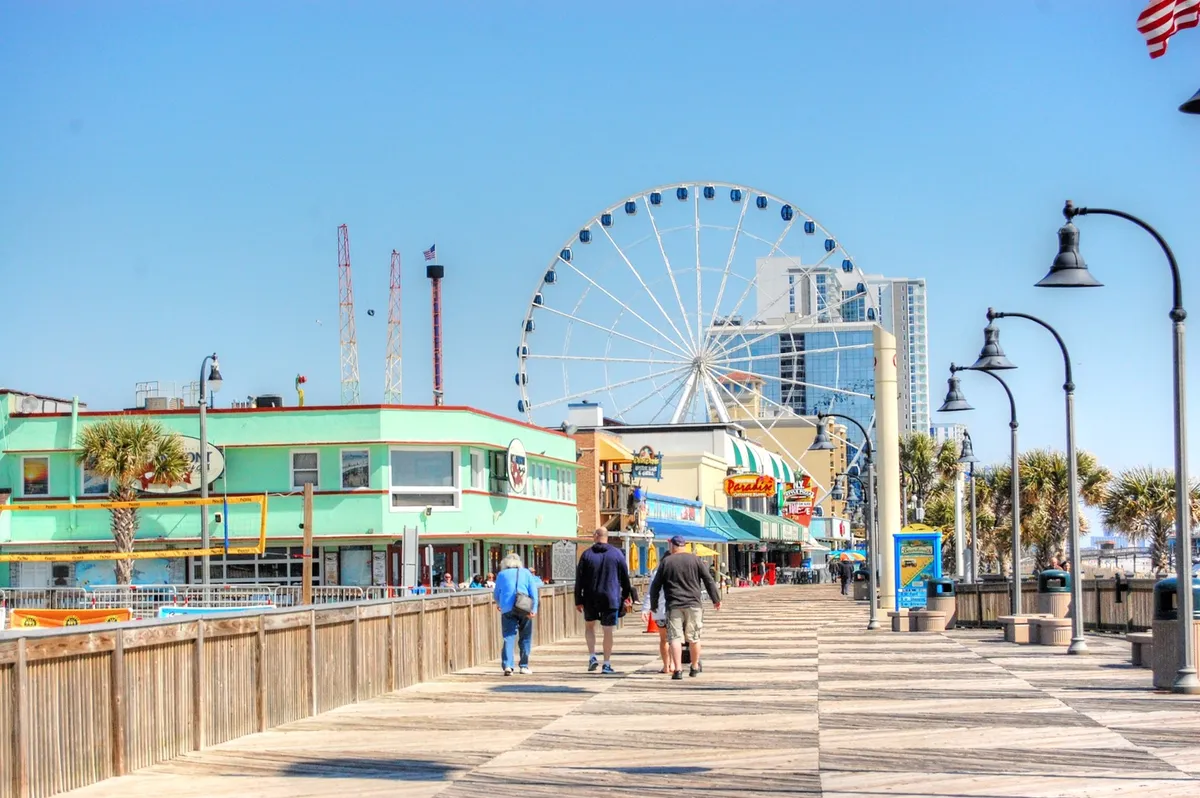 A dynamic exterior image of the local boardwalk, with a ferris wheel, colorful buildings, and people strolling. The scene bursts with energy, highlighting a sense of community fun, local dining, and entertainment all within a lively beachside setting.