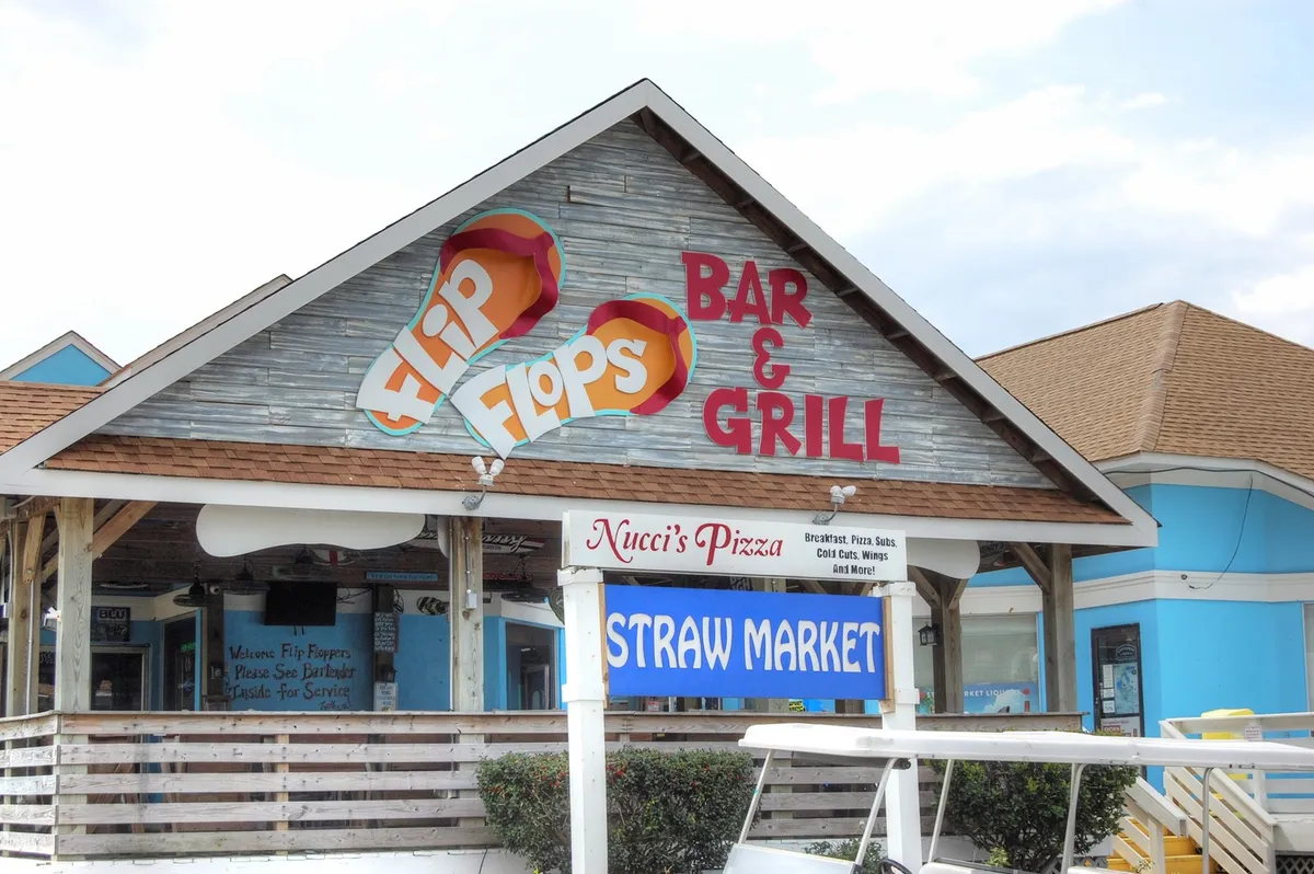 A lively exterior shot showcases the signage for local dining options like Flip Flops Bar & Grill and Nucci's Pizza, with inviting seating and an atmosphere that hints at social gatherings and live music. The scene is vibrant yet relaxed, suggesting easy access to local attractions.