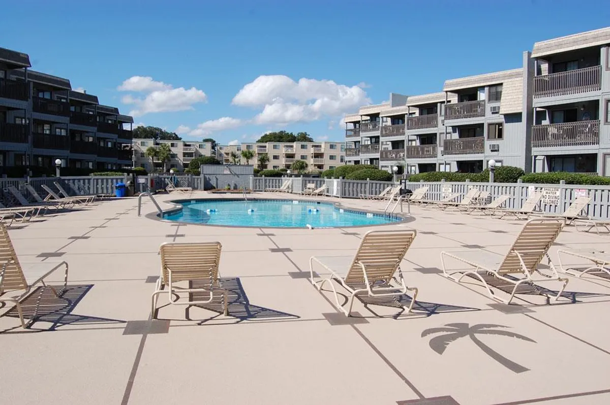A sparkling swimming pool surrounded by lounge chairs and flanked by the condo buildings. The setting is bright and inviting, with ample space for guests to relax, socialize, or enjoy a swim. The presence of multiple loungers and the pool itself emphasizes the resort-style atmosphere.