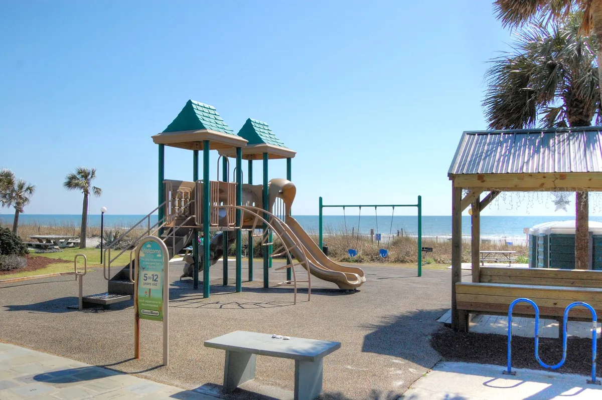 This image features a vibrant playground with swings and play equipment set just steps from the sandy beach. In the background, the beach stretches out, blending outdoor play with the classic allure of the shore, while picnic tables invite family gatherings.