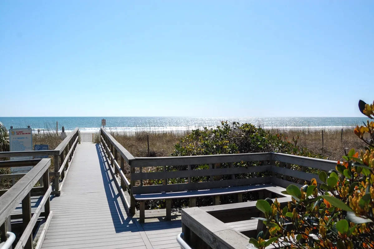 A boardwalk extends through dunes and beach grass, leading directly to the sandy beach and ocean beyond. The scene promises easy and scenic access to the shore, inviting residents to enjoy the coastline just moments from their condo.