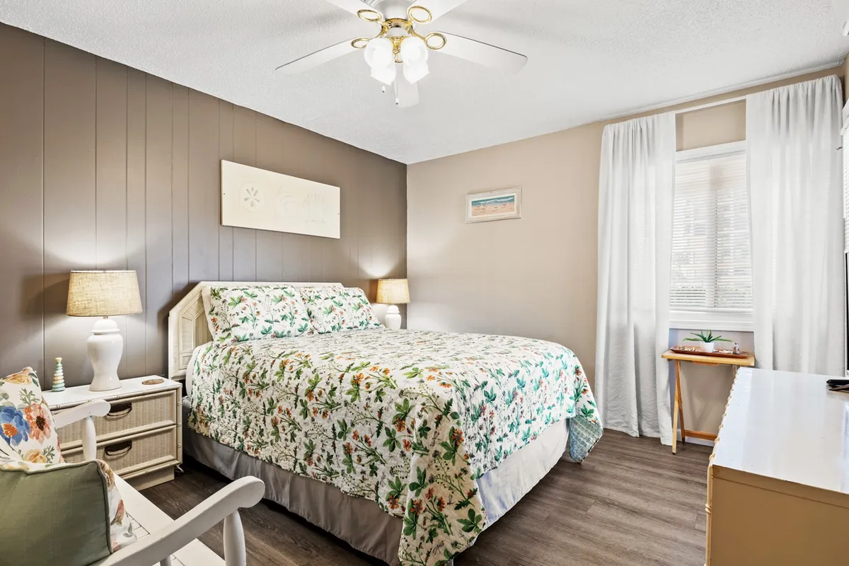 A cozy bedroom scene showcasing a plush queen bed with floral bedding, a comfortable chair with decorative pillows, a nightstand with a lamp, and a window dressed in light curtains. The space is bathed in gentle morning light, evoking relaxation and comfort.