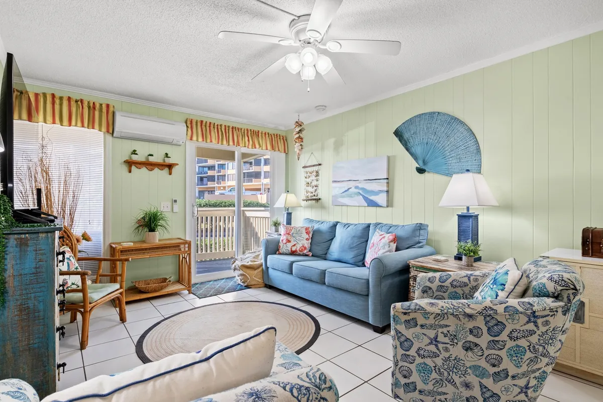 The image showcases a bright, inviting living space with a plush blue sofa, a whimsical seashell chair, coastal-themed artwork, and a dining table set for gatherings. Large windows (not visible, but suggested by the lighting and activities) hint at ocean views and create a cozy, light-filled atmosphere perfect for relaxing or socializing.